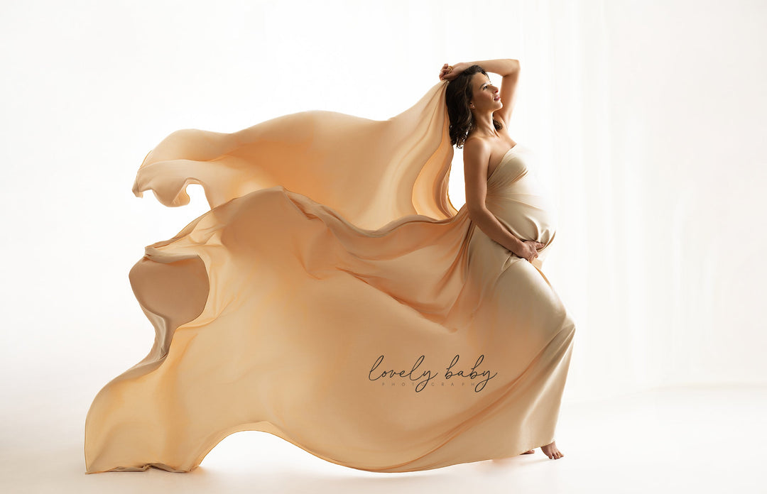 A pregnant woman in a flowing champagne-colored dress poses with a large, billowing sheer fabric drape against a bright white studio background. The dress features a script logo reading &